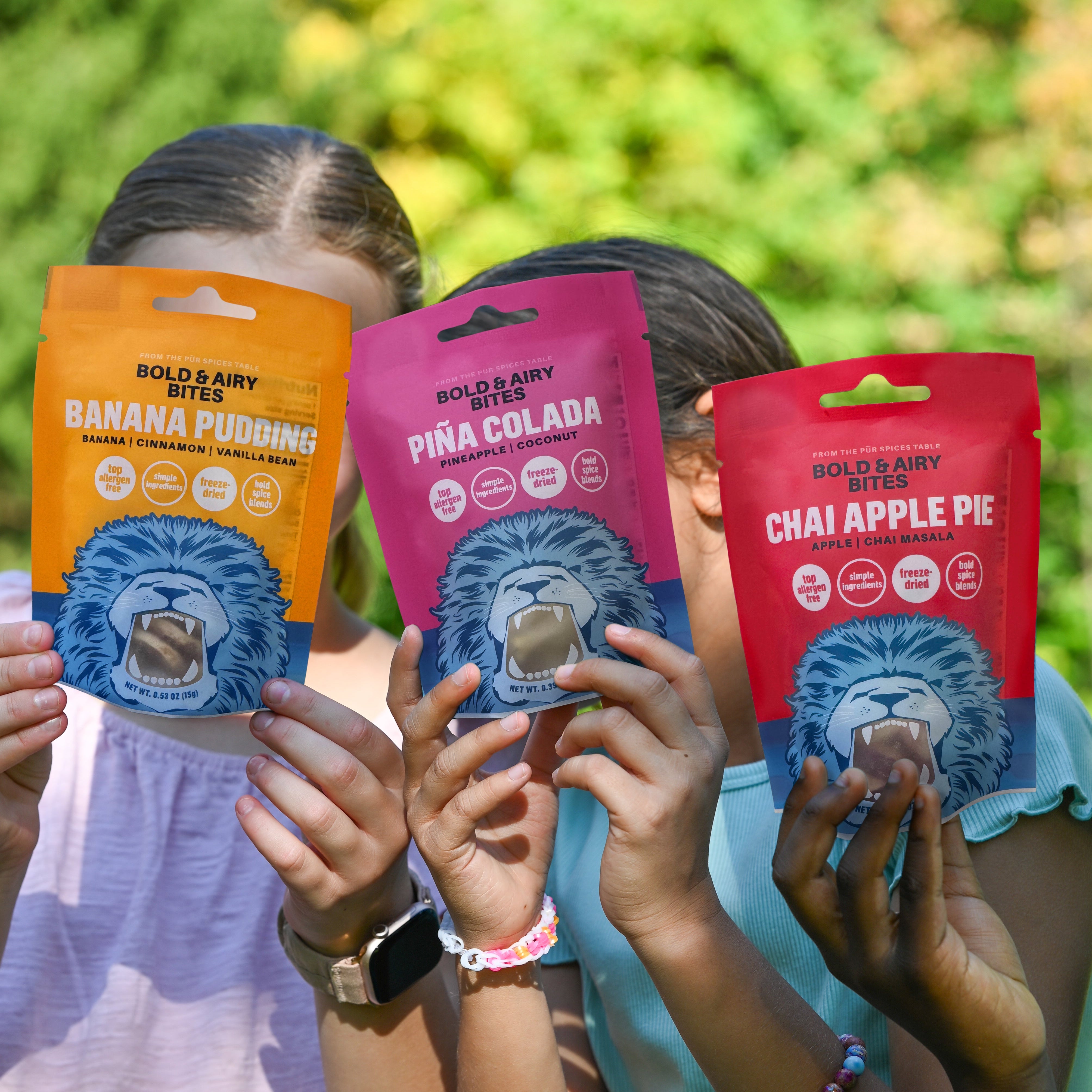 Three children outdoors holding up colorful pouches of Bold & Airy Bites. The packages are orange, pink, and red, labeled Banana Pudding, Pina Colada, and Chai Apple Pie, with a blue lion graphic on each.